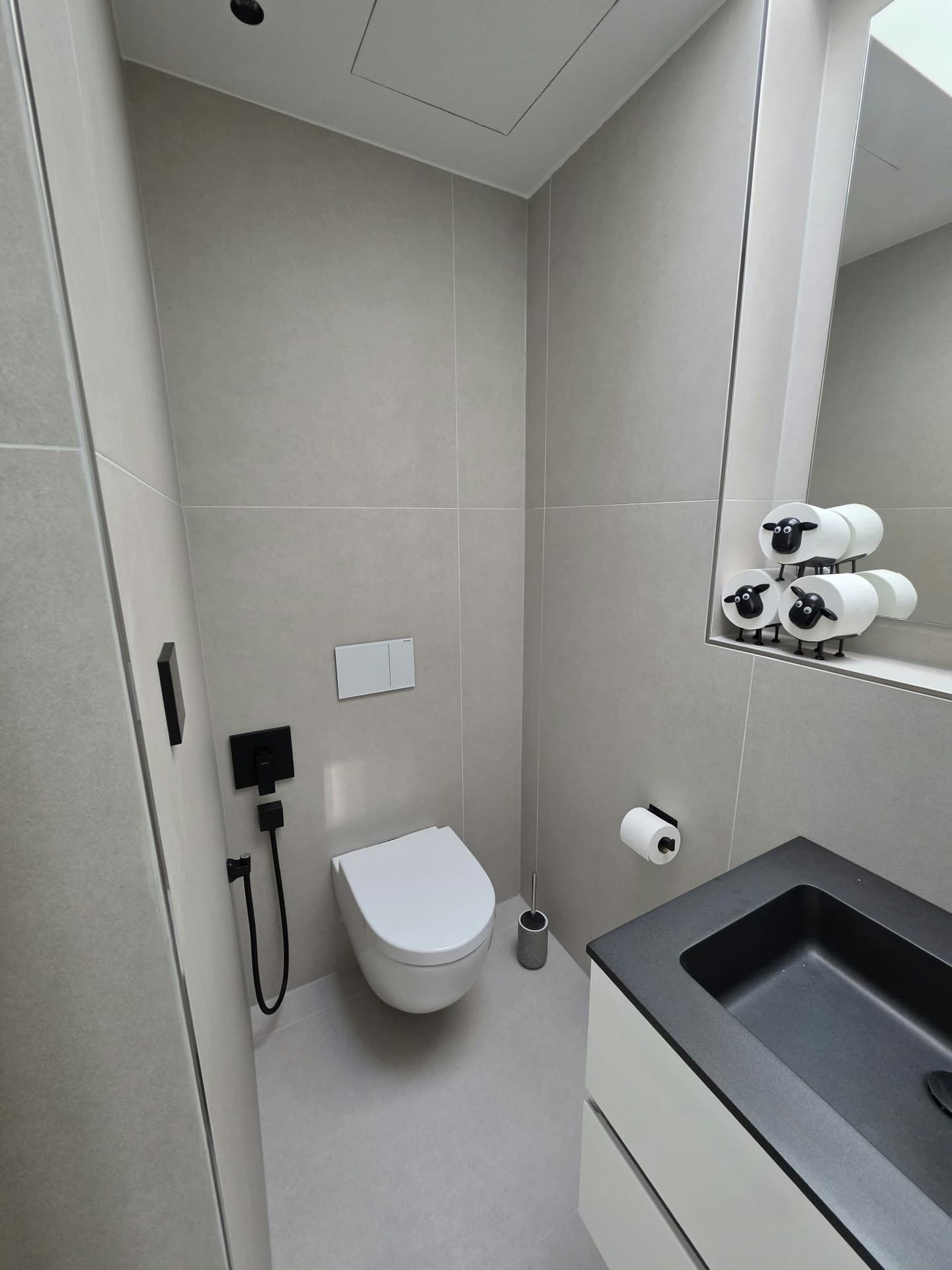 Gäste-WC mit großformatigen hellgrauen Fliesen an Wand und Boden, Wand-WC und Waschtisch mit schwarzem Becken